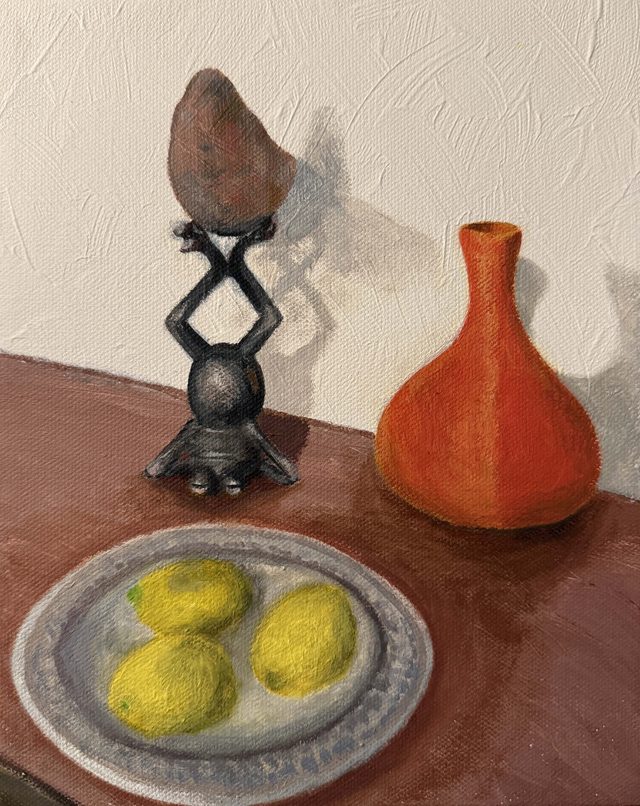 Still life of a sweet potato balanced on a small metal stand beside an orange vase and a dish of lemons