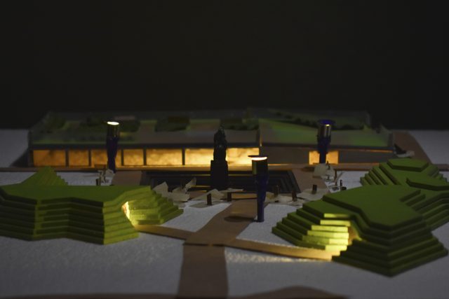 Architectural model of the proposed cultural center at night, its glowing interior set into stepped green terraces around the Masaryk statue