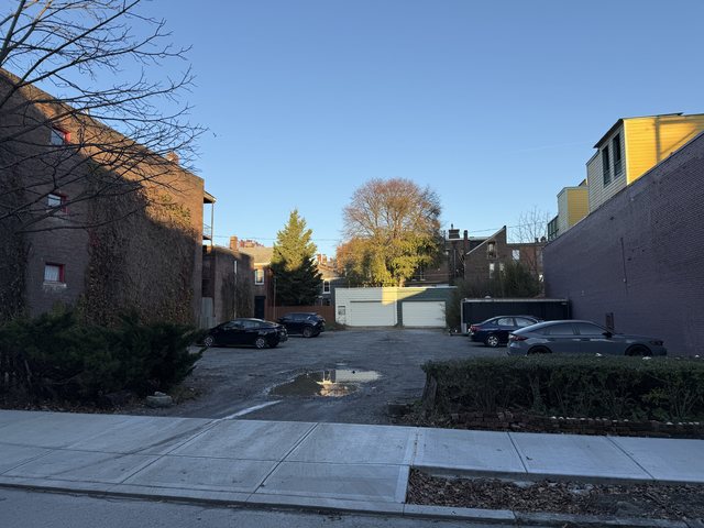 The site as it stands, a parking lot off an alley behind brick and painted buildings under a clear autumn sky